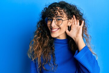 Young hispanic girl wearing casual clothes and glasses smiling with hand over ear listening and hearing to rumor or gossip. deafness concept.