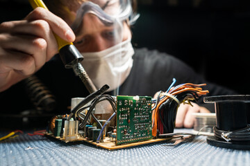 A man in glasses and a mask, with a soldering iron, repairs equipment