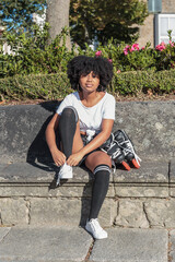 Young African American woman with afro hair looks at camera and is sitting to put on in-line roller skates to train outdoor skating in a park and play sports on hot sunny day