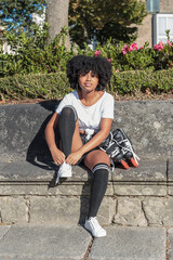 Young African American woman with afro hair looks at camera and is sitting to put on in-line roller skates to train outdoor skating in a park and play sports on hot sunny day © Graphic PhotoArt