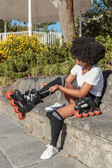 Young African American woman with afro hair is sitting putting on inline roller skates for outdoor skating training in park and playing sports on hot sunny day