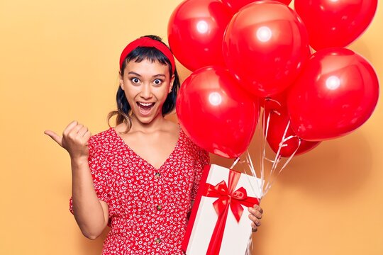 Young woman holding balloons with gift pointing thumb up to the side smiling happy with open mouth