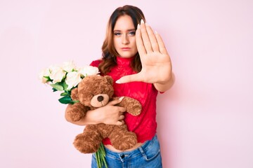 Young beautiful caucasian woman holding bear and bouquet of flowers for anniversary with open hand doing stop sign with serious and confident expression, defense gesture