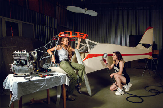 Two young women painting a body plane in airplane hangar near the table with engine and tools. Beautiful female workers in small aviation. Advertising image of aircraft making or maintenance.
