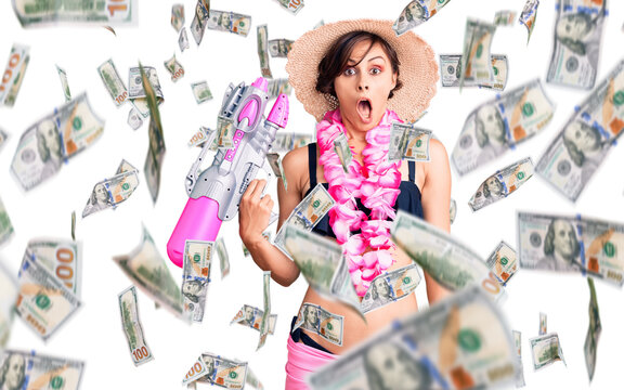 Beautiful young woman with short hair wearing bikini and hawaiian lei holding water gun scared and amazed with open mouth for surprise, disbelief face
