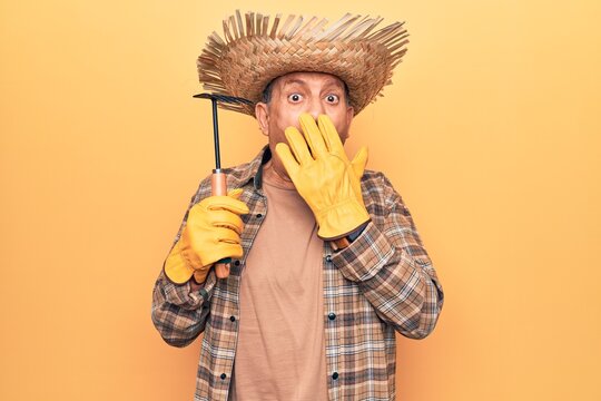 Senior man with grey hair wearing gardener hat holding rake covering mouth with hand, shocked and afraid for mistake. surprised expression