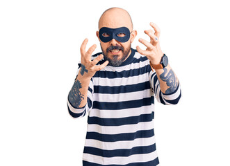Young handsome man wearing burglar mask shouting frustrated with rage, hands trying to strangle, yelling mad