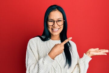 Beautiful hispanic woman wearing casual sweater and glasses amazed and smiling to the camera while presenting with hand and pointing with finger.