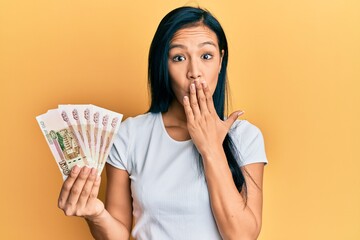 Beautiful hispanic woman holding 100 russian ruble banknotes covering mouth with hand, shocked and afraid for mistake. surprised expression