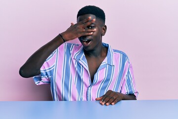 Young african american man wearing casual clothes sitting on the table peeking in shock covering face and eyes with hand, looking through fingers with embarrassed expression.