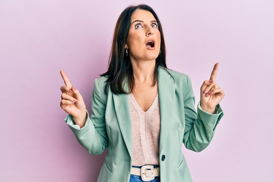 Middle age brunette woman wearing casual clothes amazed and surprised looking up and pointing with fingers and raised arms.