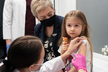 Obraz na płótnie Canvas A nurse sticks a needle into the girl's arm with the new vaccine. Grandma cheers her granddaughter during the vaccination. The doctor makes the necessary injection into the child's arm. Preventive