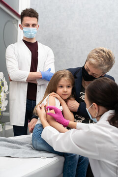 Grandma Cheers Her Granddaughter During The Vaccination. A Young Nurse Stabs A Needle Into The Girl's Arm. A Trainee In The Last Year Of Medicine Observes The Whole Situation In The Doctor's Office