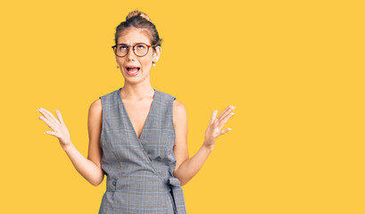 Beautiful caucasian woman with blonde hair wearing business clothes and glasses crazy and mad shouting and yelling with aggressive expression and arms raised. frustration concept.