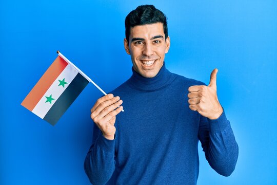 Handsome hispanic man holding syria flag smiling happy and positive, thumb up doing excellent and approval sign