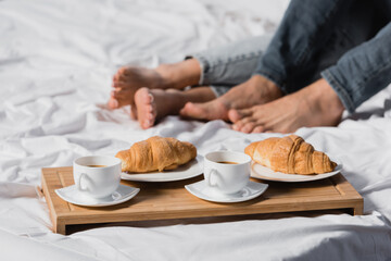 Obraz na płótnie Canvas Copped view of coffee and croissants on breakfast tray near legs of couple on blurred background on bed