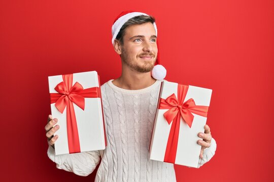 Handsome caucasian man wearing christmas hat and holding gifts smiling looking to the side and staring away thinking.