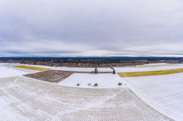Wide winter aerial view over snowy fields with trees and a cloudy horizon, shot by a drone.