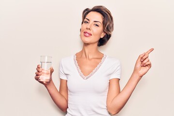 Young beautiful woman drinking glass of water smiling happy pointing with hand and finger to the side