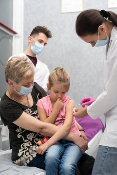 A Nurse Gives A Vaccine To A Girl Who Is Sitting On Her Grandmother's Lap Because She Feels So Safe. A Trainee In The Last Year Of Medicine Observes The Whole Situation In The Doctor's Office And