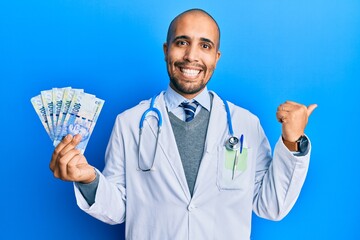 Hispanic adult doctor man wearing medical uniform holding 100 south african rand pointing thumb up to the side smiling happy with open mouth