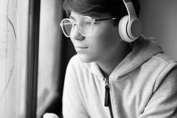 Black and white portrait of young boy. Isolation at home for self quarantine. Youngster guy spending free time home. Teenager in white headphones listening music and looking through the window.