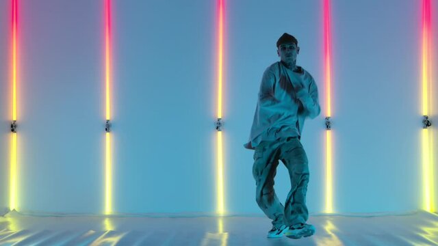 Cool young talented hip hop guy is dancing against the backdrop of bright neon lights, performing amazing gestures and movements. Street dance. Slow motion.