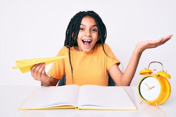 Young african american girl child with braids holding paper airplane while studying celebrating victory with happy smile and winner expression with raised hands