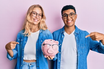 Young interracial couple holding piggy bank with glasses smiling happy pointing with hand and finger