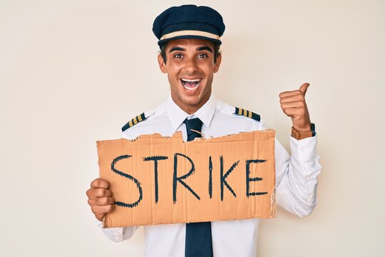 Young hispanic man wearing airplane pilot uniform holding strike banner pointing thumb up to the side smiling happy with open mouth