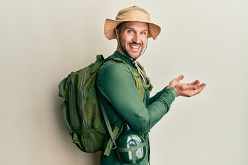 Fototapeta premium Handsome man with beard wearing explorer hat and backpack pointing aside with hands open palms showing copy space, presenting advertisement smiling excited happy