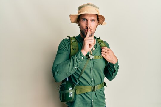 Handsome man with beard wearing explorer hat and backpack asking to be quiet with finger on lips. silence and secret concept.
