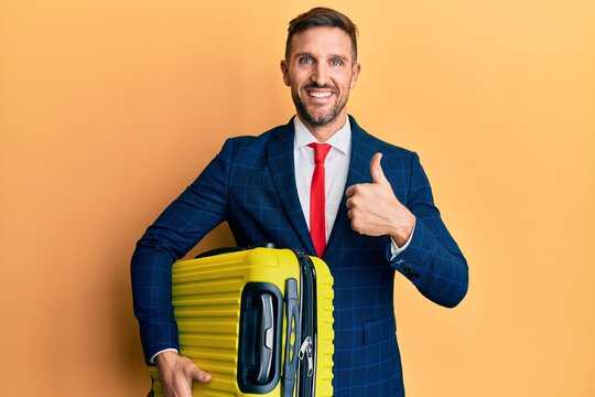 Handsome man with beard wearing business suit holding suitcase going on work trip smiling happy and positive, thumb up doing excellent and approval sign
