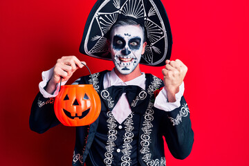 Young man wearing mexican day of the dead costume holding pumpkin screaming proud, celebrating victory and success very excited with raised arms
