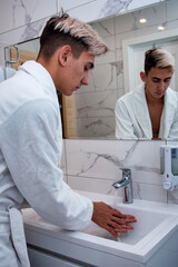 Morning procedures in the bathroom. A young man in a white coat stands at the mirror. Problem skin and care for it. Non-standard appearance.