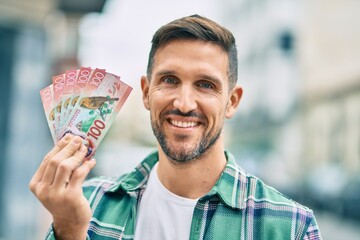 Young caucasian man smiling happy standing holding new zealand dollars banknotes at the city.