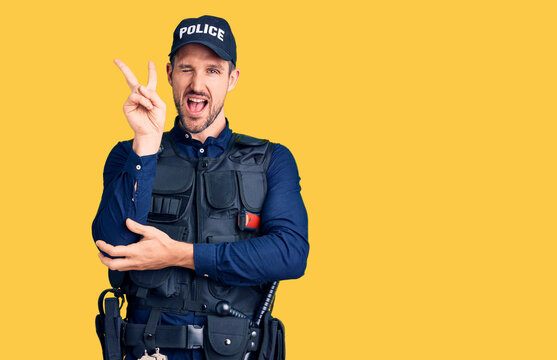 Young handsome man wearing police uniform smiling with happy face winking at the camera doing victory sign with fingers. number two.