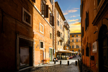 A narrow back alley in the historic center of Rome, Italy, leads to a small sidewalk cafe at sunset.