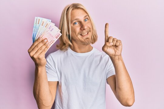 Caucasian young man with long hair holding new taiwan dollars banknotes smiling with an idea or question pointing finger with happy face, number one