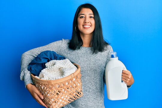 Beautiful asian young woman holding laundry basket and detergent bottle smiling with a happy and cool smile on face. showing teeth.
