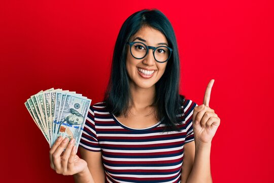 Beautiful asian young woman holding dollars smiling with an idea or question pointing finger with happy face, number one