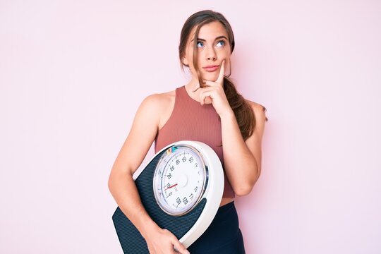 Beautiful young caucasian woman holding weight machine to balance weight loss serious face thinking about question with hand on chin, thoughtful about confusing idea