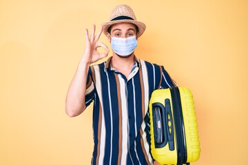 Young handsome man wearing medical mask holding suitcase doing ok sign with fingers, smiling friendly gesturing excellent symbol