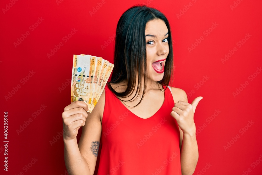 Poster young hispanic girl holding 500 philippine peso banknotes pointing thumb up to the side smiling happ