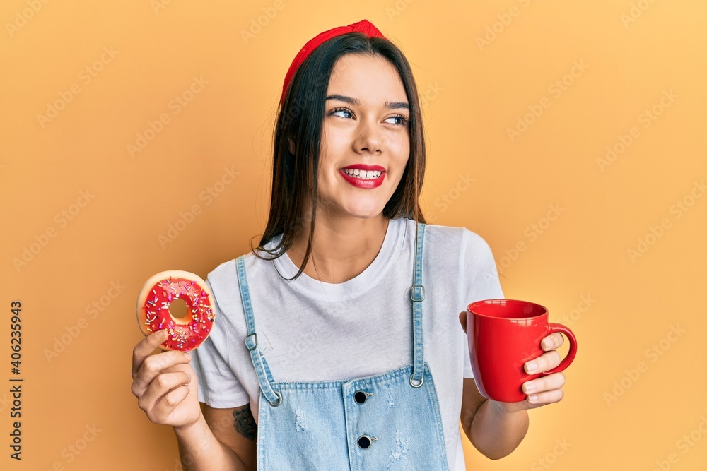 Wall mural Young hispanic girl eating doughnut and drinking coffee smiling looking to the side and staring away thinking.