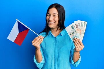 Young latin woman holding czech republic flag and koruna banknotes winking looking at the camera...