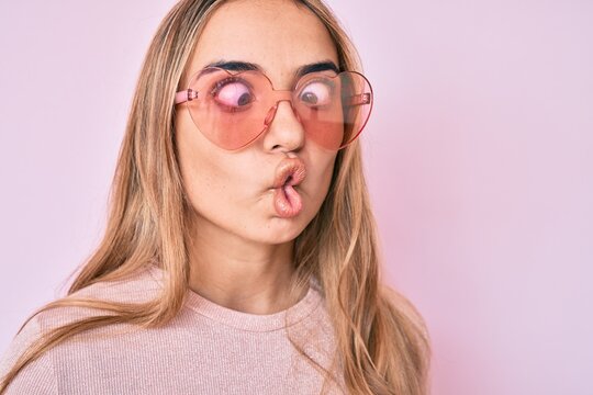 Young beautiful blonde woman wearing heart shaped sunglasses making fish face with mouth and squinting eyes, crazy and comical.