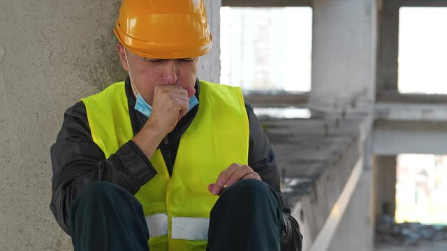 Builder in medical protective face mask and hard hat sits at construction site. He coughs and he is not feeling well