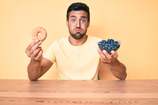 Young Hispanic Man Eating Breakfast Holding Donut And Blueberries Puffing Cheeks With Funny Face. Mouth Inflated With Air, Catching Air.