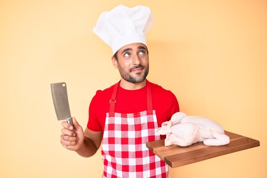 Young Hispanic Man Cooking Chicken Holding Knife Smiling Looking To The Side And Staring Away Thinking.